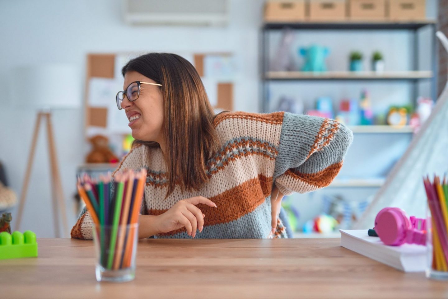 Young beautiful teacher woman wearing sweater and glasses sitting on desk at kindergarten Suffering of backache, touching back with hand, muscular pain